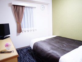 Una camera d'albergo con un letto, una scrivania con televisore, una sveglia e una finestra con tende.