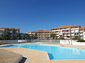 A swimming pool area with a clear blue sky, surrounded by multi-story residential buildings with balconies and palm trees.