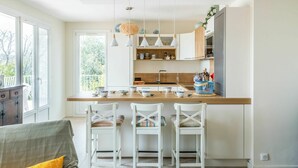 A modern kitchen with a central island, white bar stools, pendant lighting, and a view of greenery outside. - Lovely apartment for 6 guests with WIFI, A/C, TV and terrace (Saint-cyr-sur-mer)