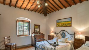 Um quarto com teto de madeira, uma cama com colcha florida, cômoda, cadeira e pintura de um campo de girassóis.