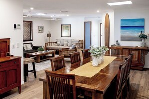 Un coin repas avec une table en bois, des chaises et un vase de fleurs. On aperçoit un canapé et une planche de surf à l’arrière-plan.