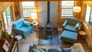 A spacious loft-style room with a high ceiling, wooden beams, a piano, two sofas, a fireplace, and large windows.