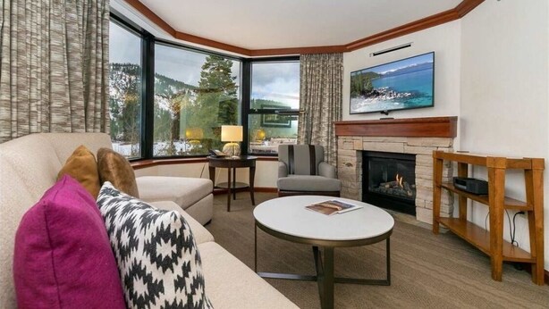 A cozy living room with a fireplace, a television, and large windows offering a scenic view.