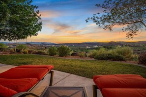 A patio with two red cushioned chairs and a table, overlooking a landscaped garden and distant hills during sunset.