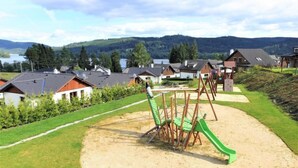 Un parque infantil con columpios y un tobogán, rodeado de casas y un lago al fondo.