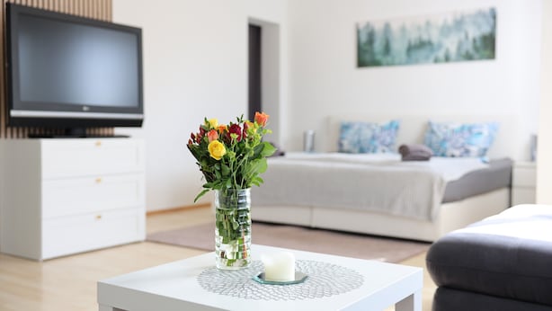 Ein modernes Wohnzimmer mit Flachbildfernseher, Blumen in einer Vase, einem weißen Couchtisch und einem Bett mit einem Gemälde an der Wand.