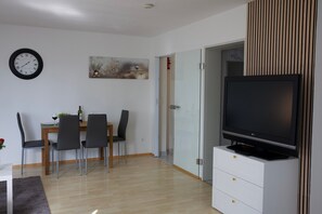 En moderne stue med spiseplads, et fladskærms-tv på et stativ, et vægur og en dør til et andet rum.