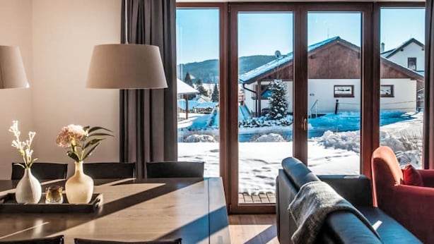Una sala de estar moderna con un amplio ventanal corredera que da a un espacio exterior nevado, una mesa de comedor con jarrones y un sofá confortable.
