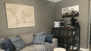 Un salon avec un canapé gris, des coussins à motifs, une carte du monde, une pancarte indiquant « Partons à l’aventure », un four à micro-ondes, une machine à café et une petite table ronde.