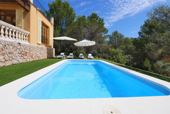 A swimming pool with lounge chairs and umbrellas, surrounded by a stone wall and greenery.