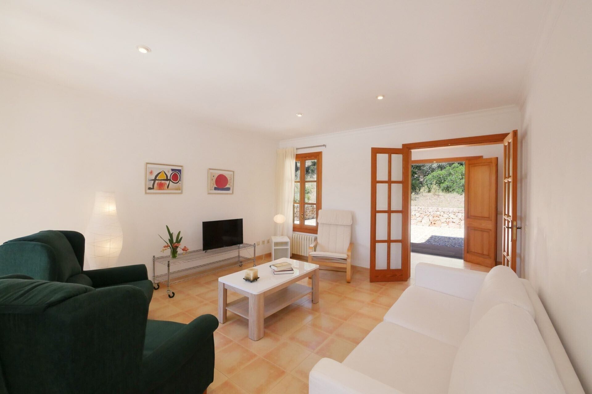 Ein Wohnzimmer mit einer Couch, einem Fernseher, einem Couchtisch und einem Sessel. An der Wand hängen zwei Gemälde und eine Tür führt nach draußen.