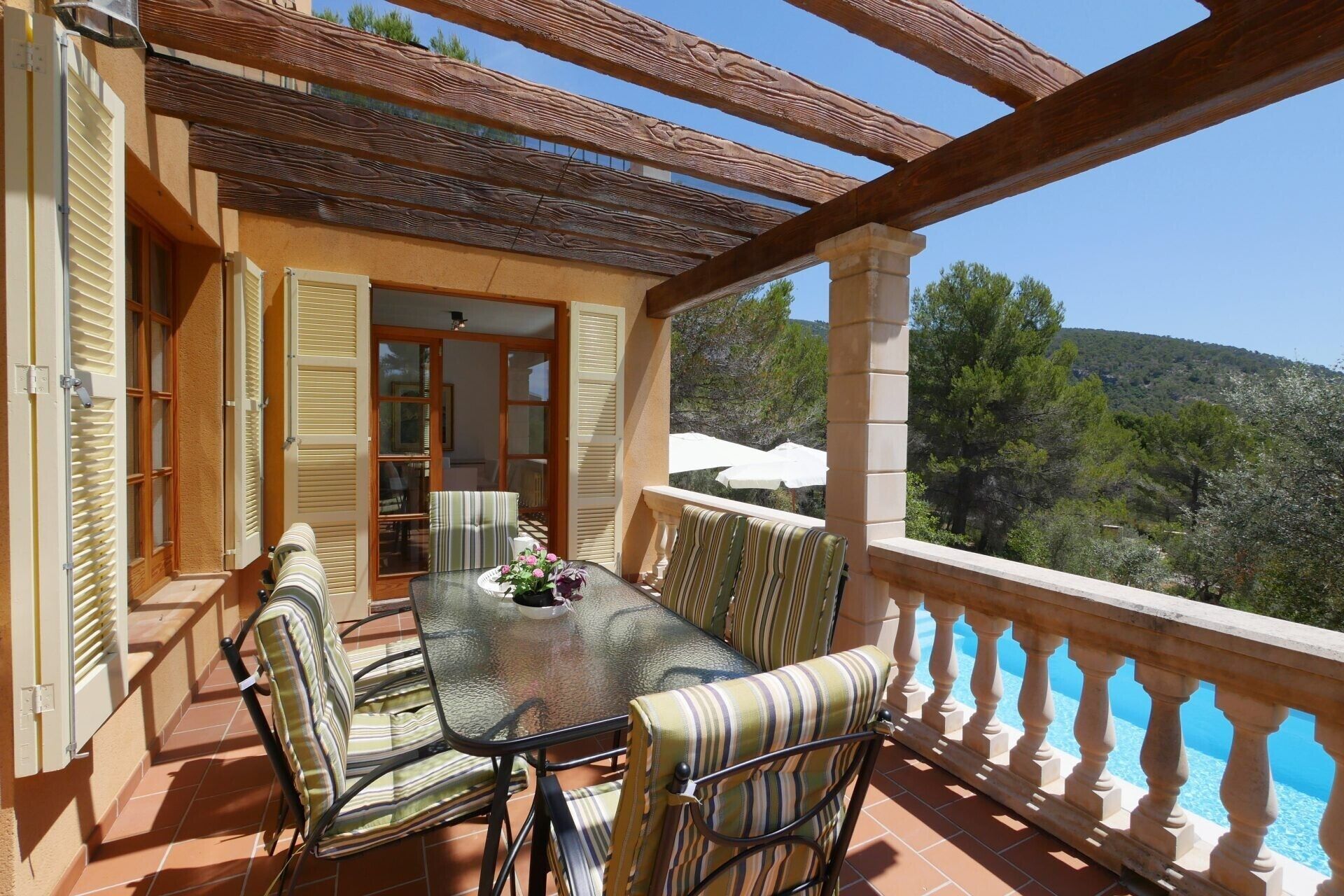 Un balcon avec une table et des chaises, une vue sur des arbres et une colline, et un bâtiment avec des volets.