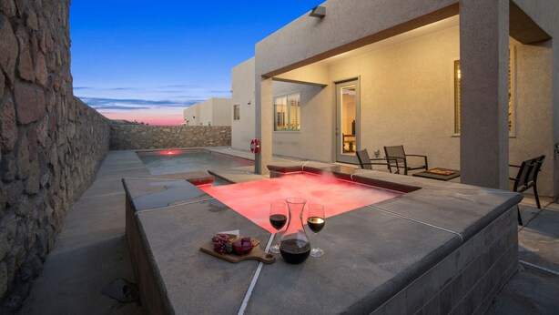 A modern outdoor pool area with a stone wall, lounge chairs, and a table with wine glasses and grapes. - Three Trees Saltwater Pool Place-Heated Pool (El Paso)
