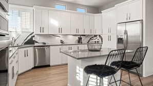A modern kitchen with white cabinets, stainless steel appliances, a marble backsplash, and a central island with two black chairs. - Three Trees Saltwater Pool Place-Heated Pool (El Paso)
