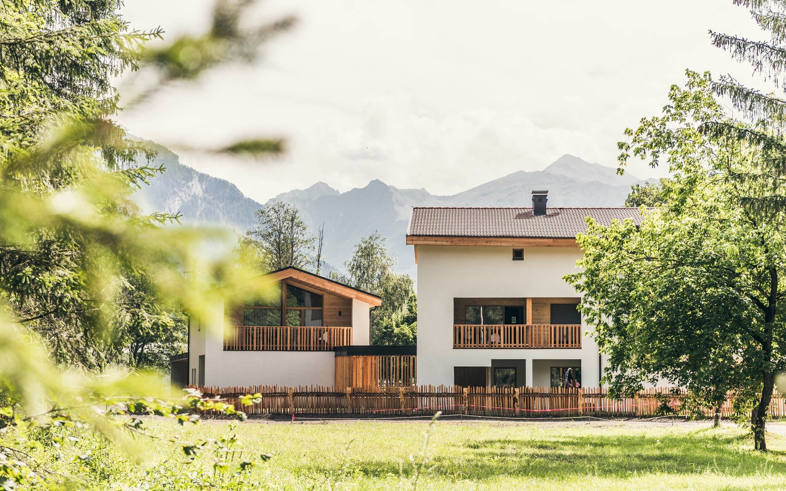 Un edificio moderno su due piani con un recinto in legno di fronte, immerso nel verde e con montagne sullo sfondo.