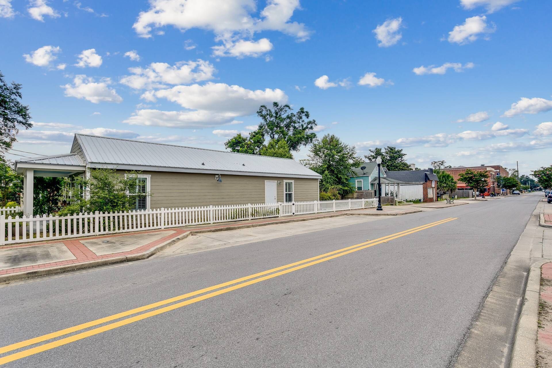 A quiet street with a single-story building, a white picket fence, and a clear sky.
