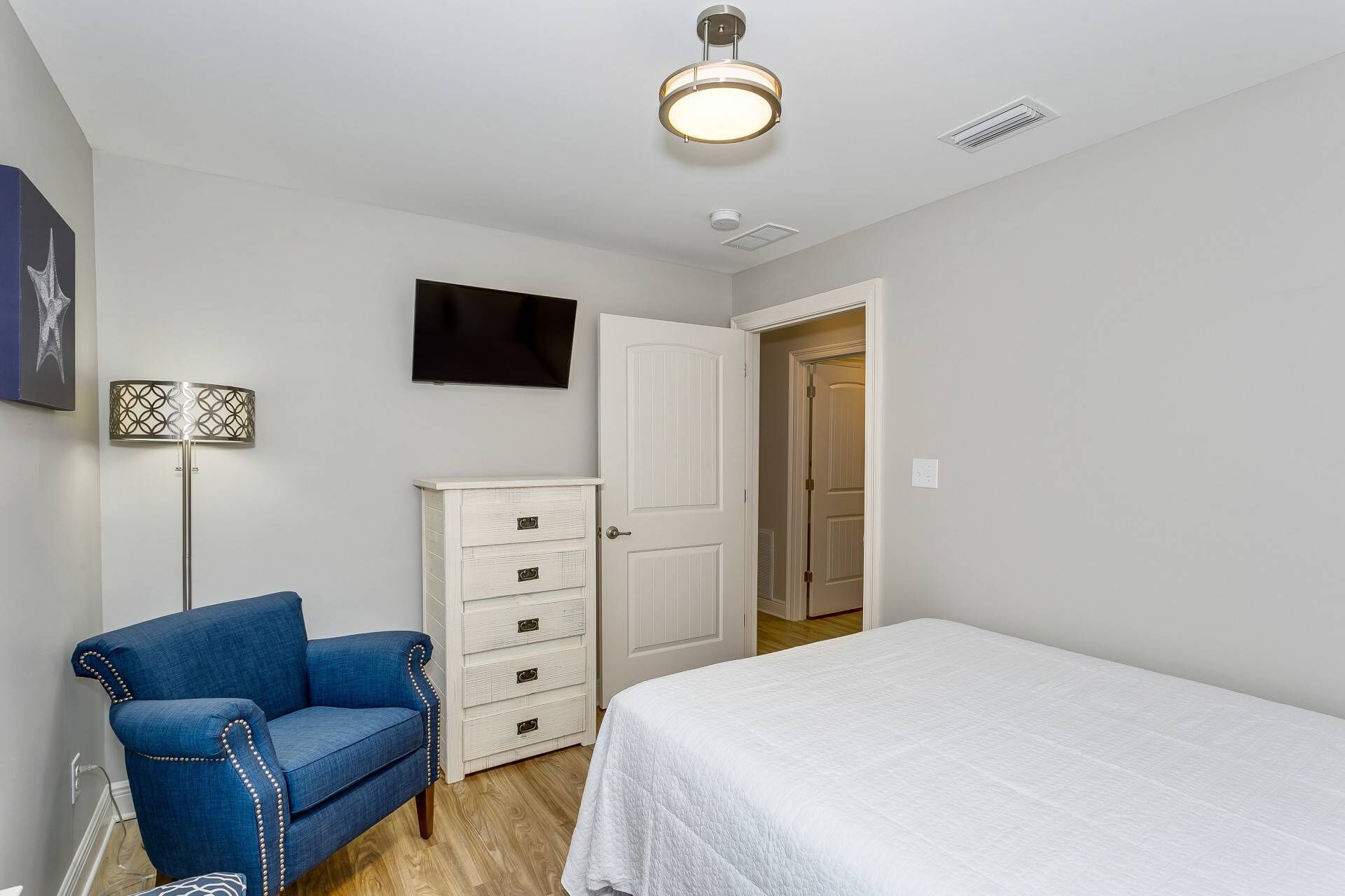 A bedroom with a bed, a blue chair, a white dresser, a floor lamp, and a TV on the wall.