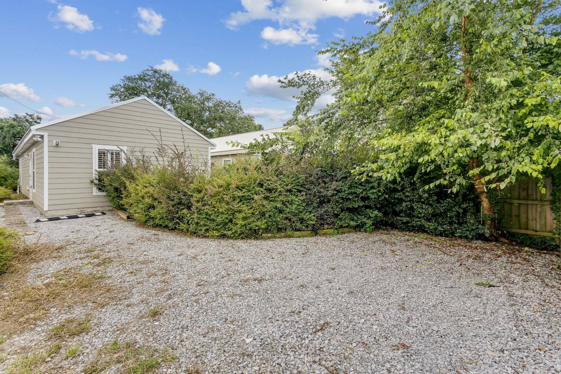 A house with a gravel driveway, surrounded by dense shrubbery and a wooden fence.