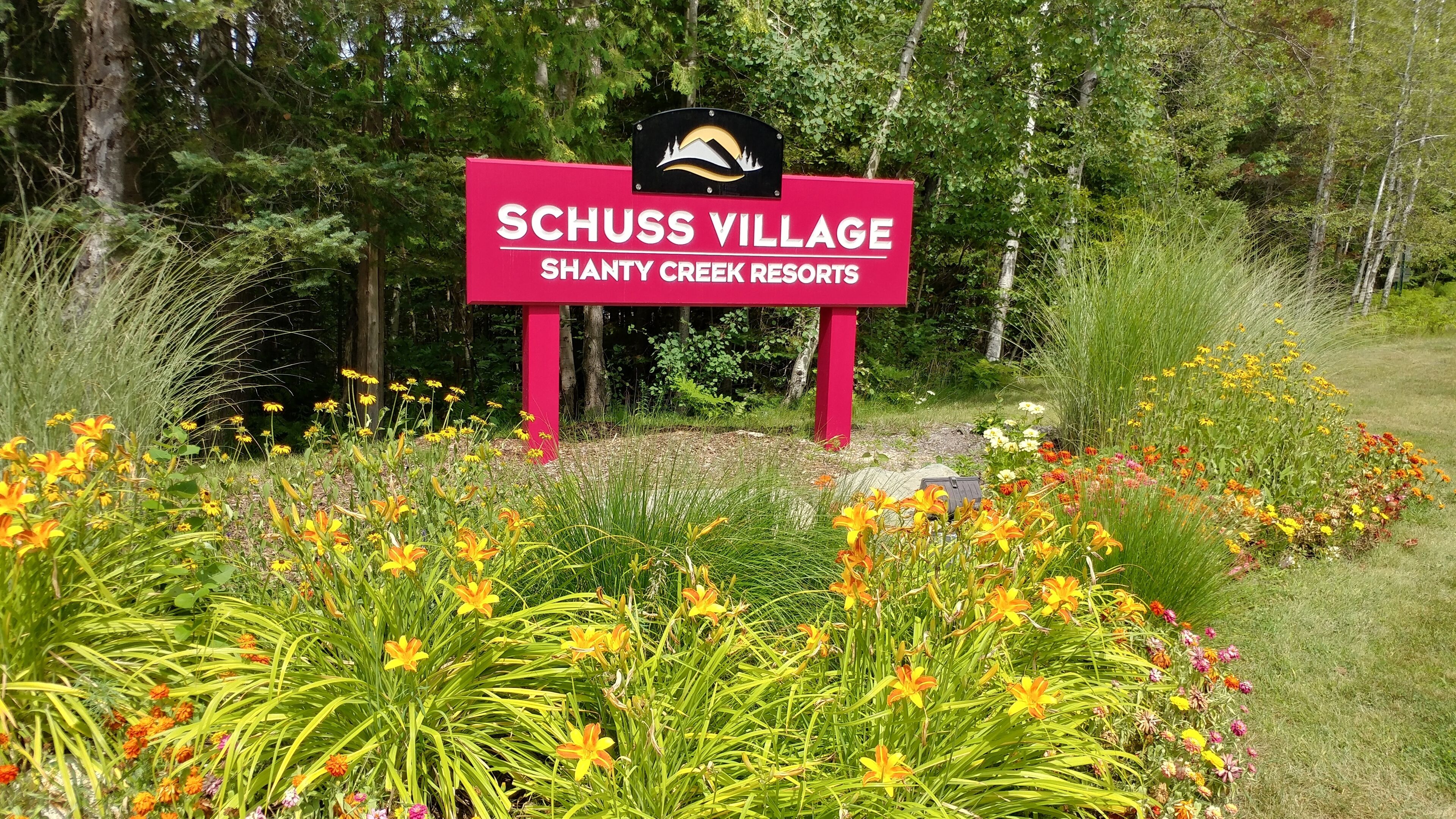 Een bordje voor Schuss Village bij Shanty Creek Resorts, omringd door een tuin met verschillende bloemen.
