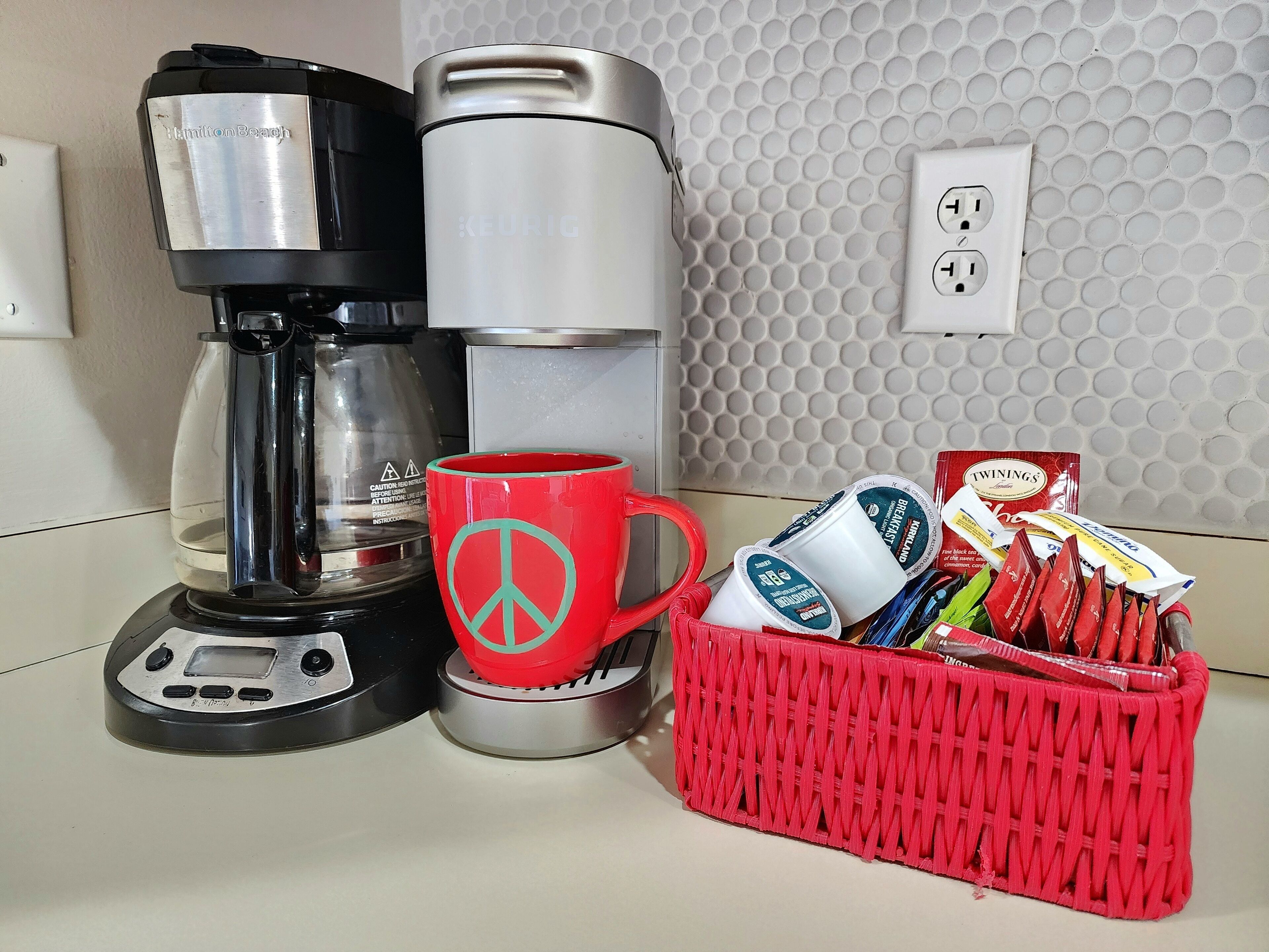 Een keukenhoek met een koffiezetapparaat, een waterdispenser, een rode mok met een vredesteken en een mandje Twinings theezakjes.