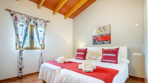 Camera d'albergo con due letti, una coperta rossa, biancheria da letto bianca, una finestra con tende, un quadro appeso al muro e un pavimento piastrellato.
