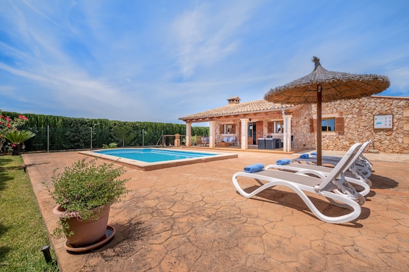 A pool area with lounge chairs, a stone building, and a pergola with a thatched roof.