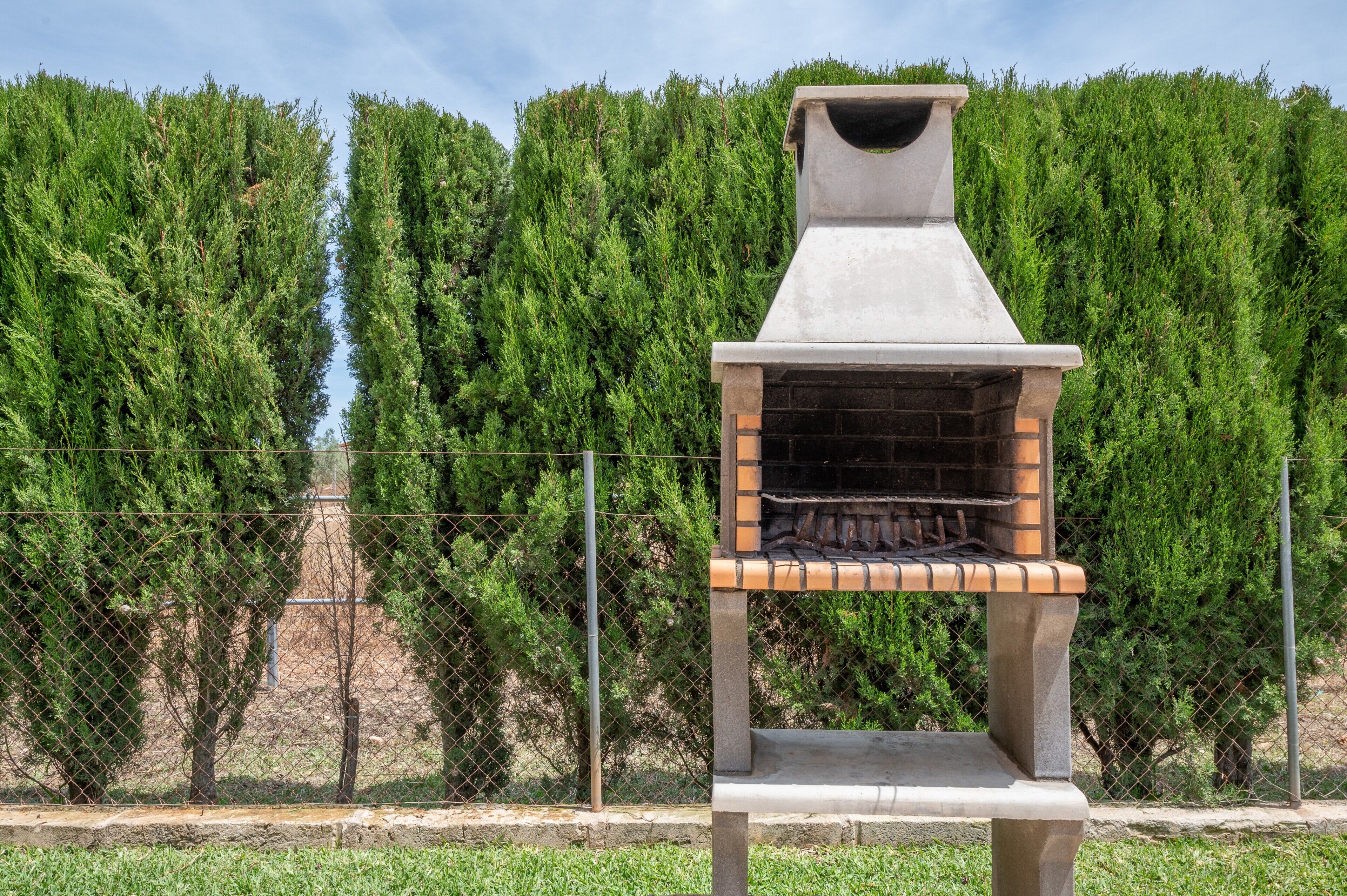 Een buitenbarbecue van bakstenen met een schoorsteen, met op de achtergrond hoge groene bomen en een gaashek.