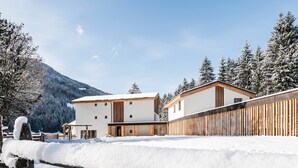 Edificio moderno con fachada blanca y detalles de madera, rodeado de árboles cubiertos de nieve y una montaña al fondo.