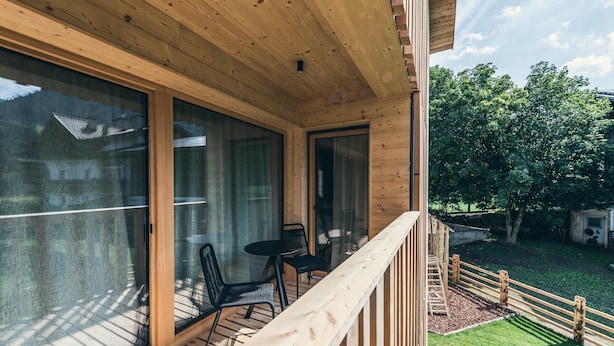Una terraza de madera con puerta de vidrio que da a una habitación, una mesita con sillas y una barandilla de madera.
