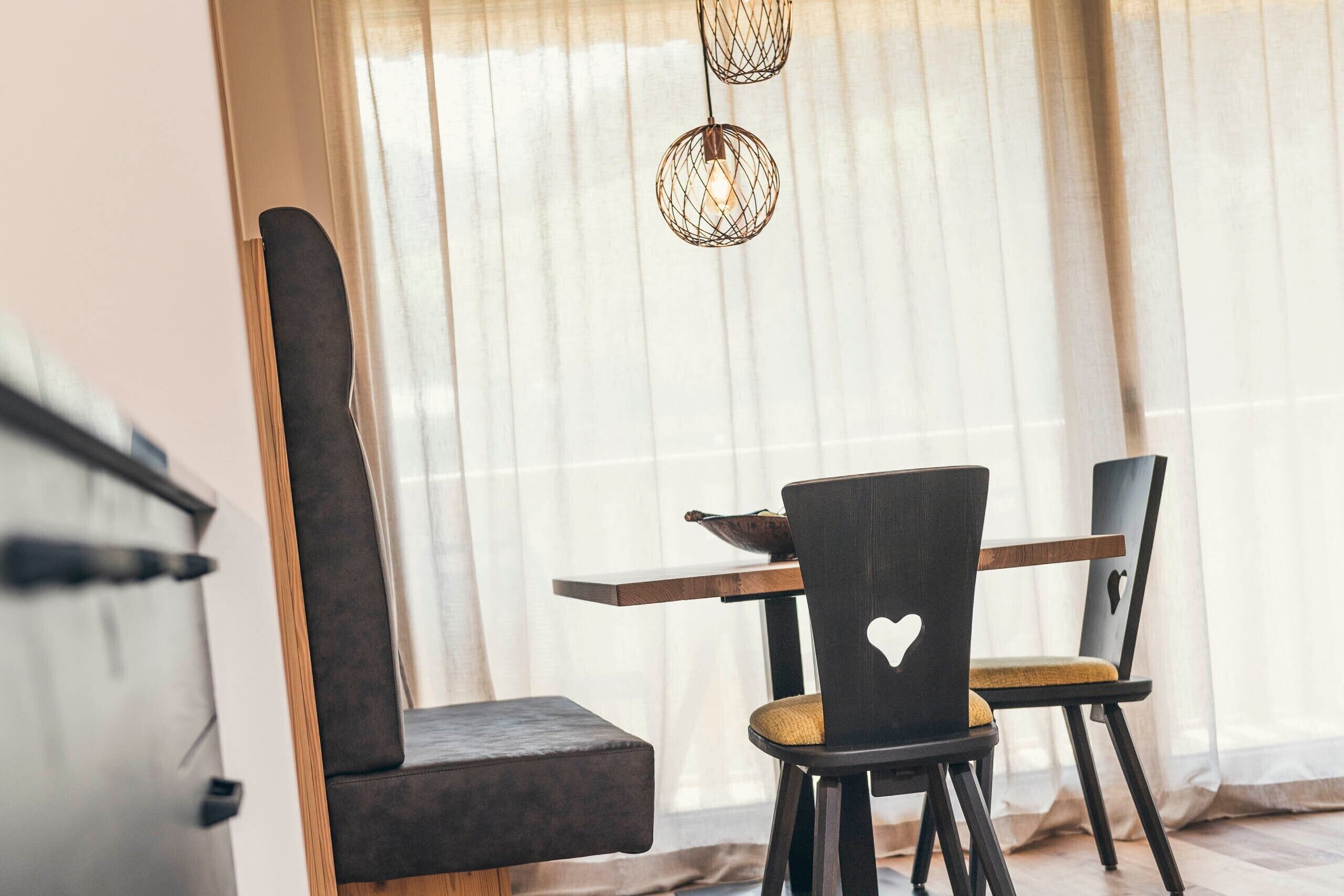 Een eethoek met een tafel en stoelen, een raam met dunne gordijnen, en een hanglamp.