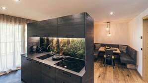 Une cuisine moderne dotée d’une cuisinière intégrée, d’un coin repas avec un banc et d’une vue sur une forêt, visible par une fenêtre.