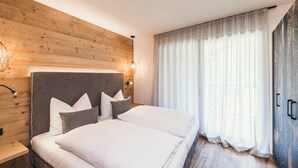Une chambre à coucher avec une tête de lit en bois, un lit recouvert de linge de lit blanc, une table de chevet et une grande fenêtre ornée de rideaux légers.