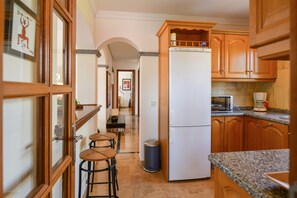 A kitchen with wooden cabinets, a refrigerator, a microwave, and a coffee maker. There are stools and a trash can.