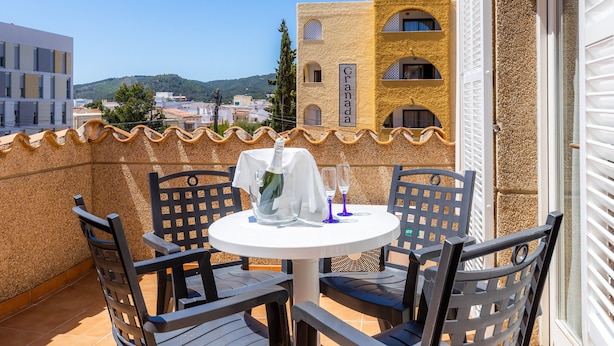 A balcony with a table set for two, a bottle of champagne, and two glasses. The balcony overlooks a cityscape with buildings and a hill in the distance.