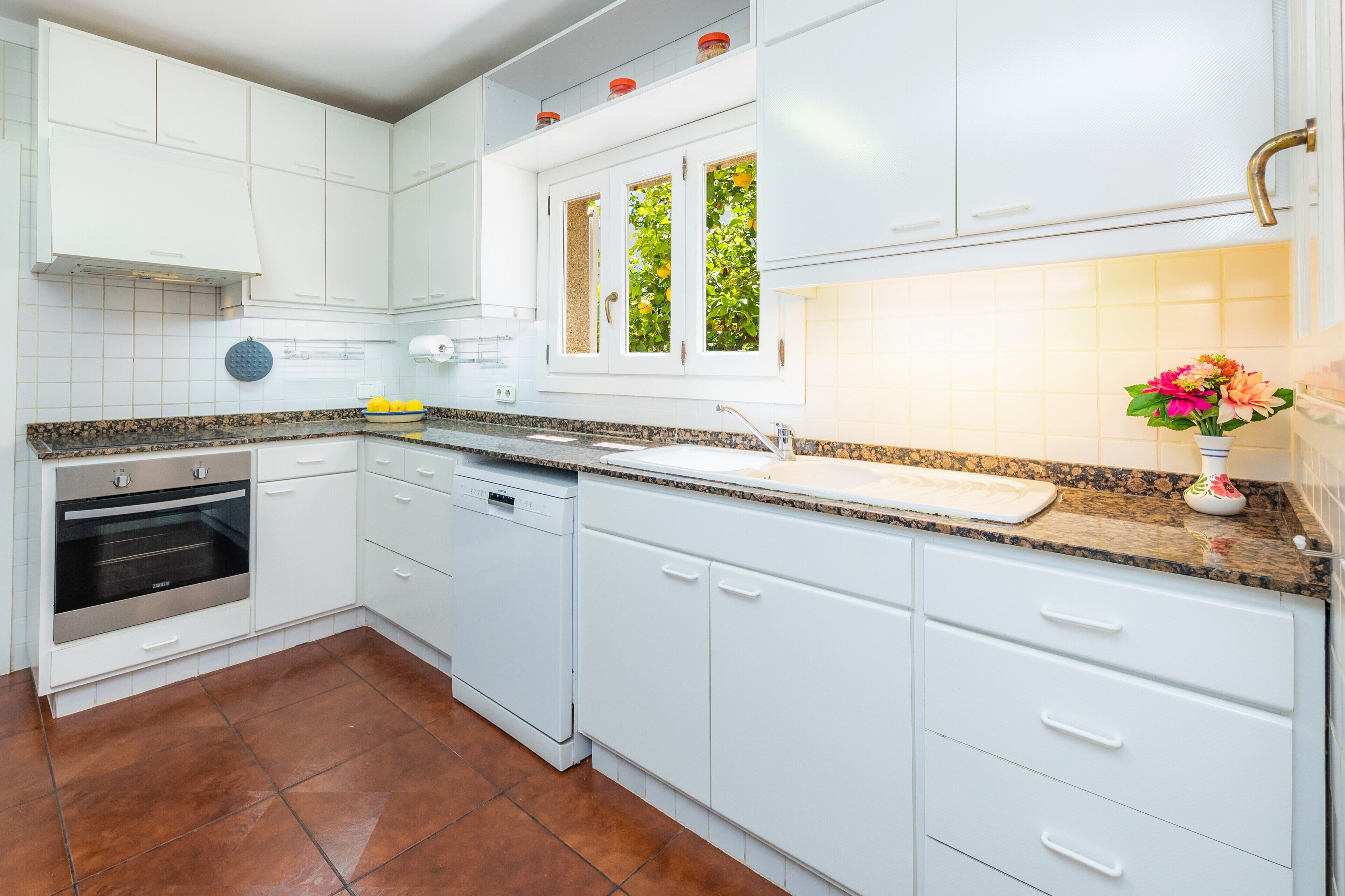 Une cuisine moderne avec des meubles blancs, un four encastré, un lave-vaisselle et une fenêtre donnant sur un espace verdoyant.