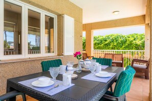 Comedor con una mesa dispuesta para cuatro, asientos exteriores, una ventana con persianas y vistas al verdor.