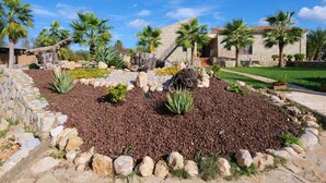 A stone building with palm trees, a garden with red gravel and rocks, and a clear blue sky.