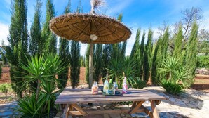 A wooden picnic table with a thatched umbrella, surrounded by greenery and stone pathways.