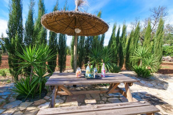 Une table de pique-nique en bois, surmontée d’un parasol en chaume, entourée de verdure et de sentiers pavés de pierres.