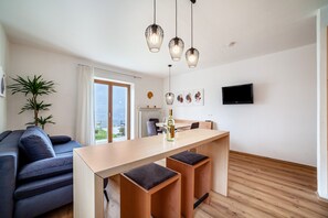 Een moderne woonruimte met een bank, een houten eettafel met krukken, hanglampen, een flatscreen televisie en een potplant.
