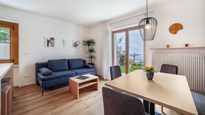 A modern living room with a sofa, dining table, chairs, a small coffee table, a hanging light fixture, a potted plant, and a view through a large window. - Apartment "Haus Hohenrain 2" with Mountain View, Pool, Wi-Fi, Terrace & Garden (Schenna)