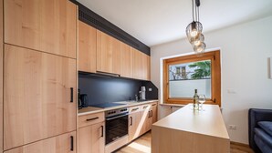 A modern kitchen with wooden cabinets, a central island, and a window with blinds. - Apartment "Haus Hohenrain 2" with Mountain View, Pool, Wi-Fi, Terrace & Garden (Schenna)