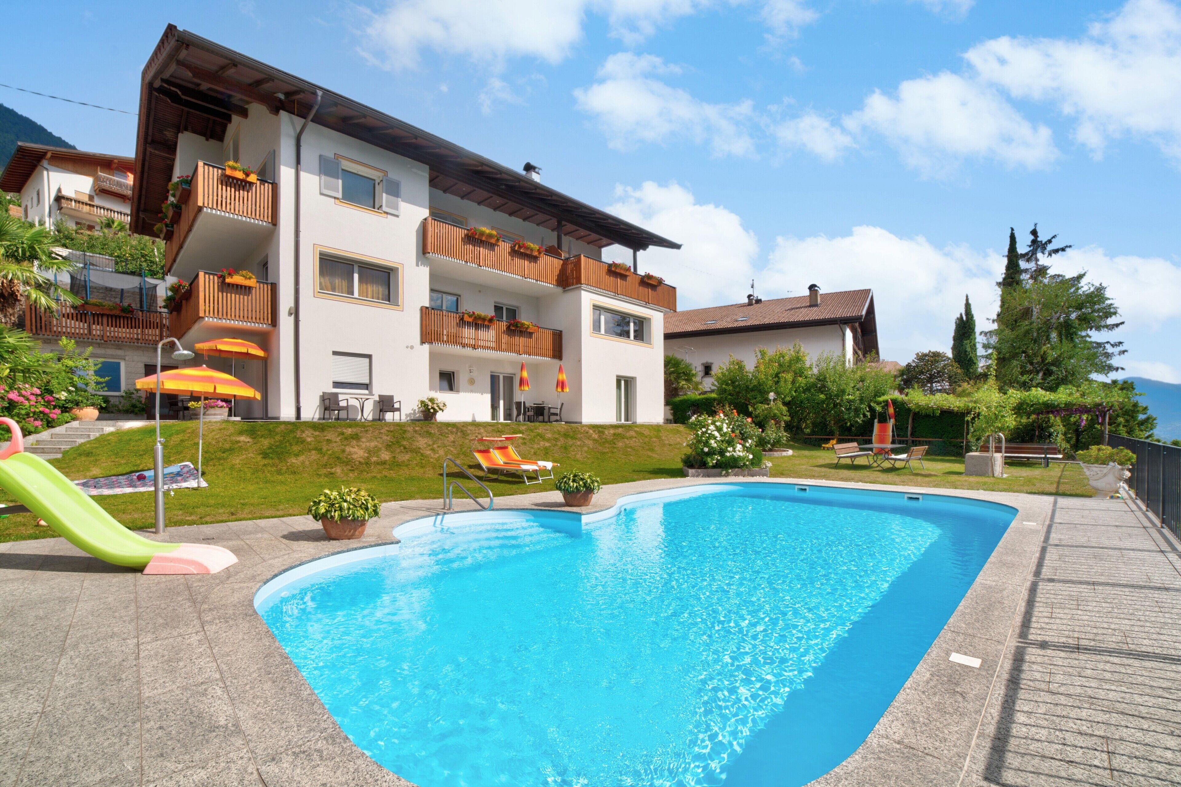 A swimming pool with a slide, surrounded by a wooden deck, and a multi-story building with balconies.