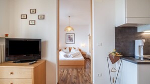 A room with a bed, a television on a wooden cabinet, a kitchen area with a coffee machine, and a hallway with framed pictures. - Apartment "Haus Hohenrain 3" with Mountain View, Pool, Wi-Fi, Terrace & Garden (Schenna)