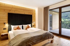 A bedroom with a wooden headboard, a large bed with white linens, a brown throw blanket, and a sliding glass door leading to a balcony with a view of a forest.