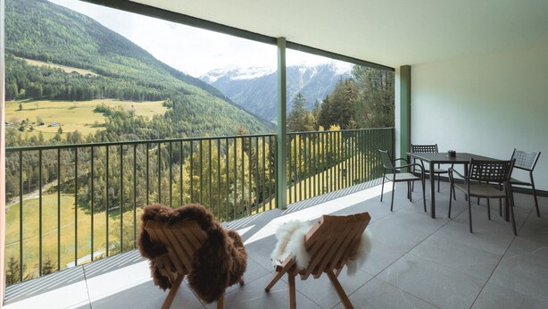 A balcony with a view of a mountainous landscape, a table with chairs, and two chairs with fur throws.