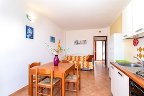 A dining area with a wooden table and chairs, a purple vase with flowers, a sofa with a patterned blanket, and a kitchen with a sink and cabinets.