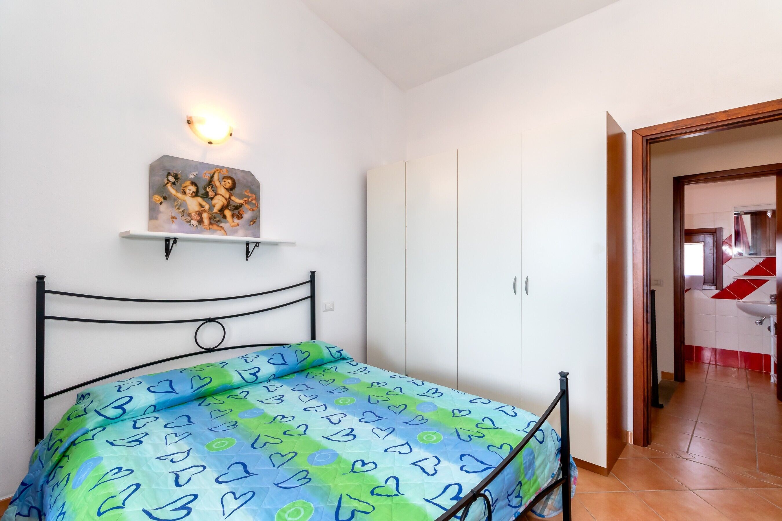 Una camera da letto con un letto, una lampada a parete, uno scaffale con un quadro e vista sul bagno.