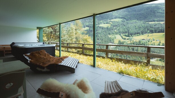 Una habitación moderna con un ventanal grande, un jacuzzi y una terraza de madera con vistas a un valle boscoso.