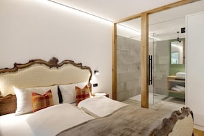 Moderni hotellihuone, jossa on suuri sänky, lasiseinällä varustettu suihku ja puinen pääty.